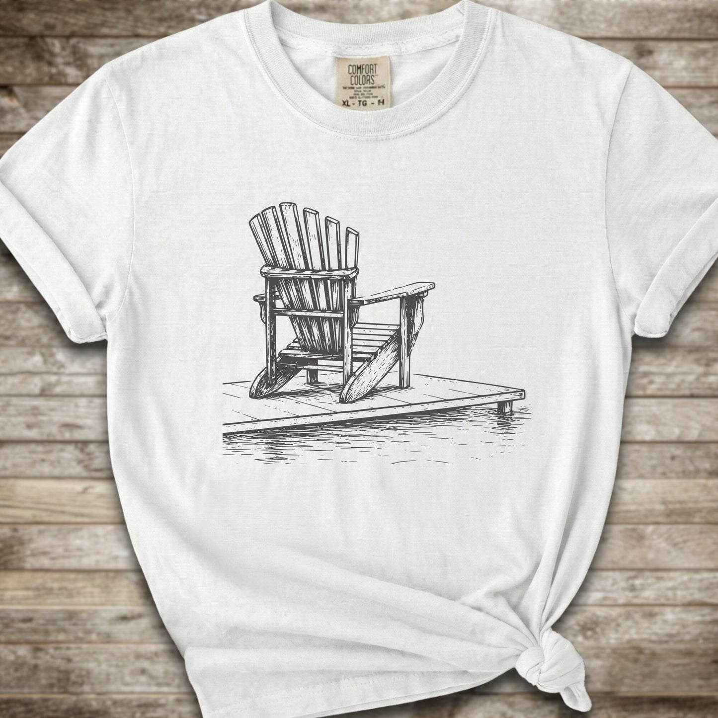 A white short-sleeve t-shirt with a white line art graphic of a Muskoka / Adirondack chair on a dock, styled with a knot at the bottom hem and laid flat on a rustic wood 