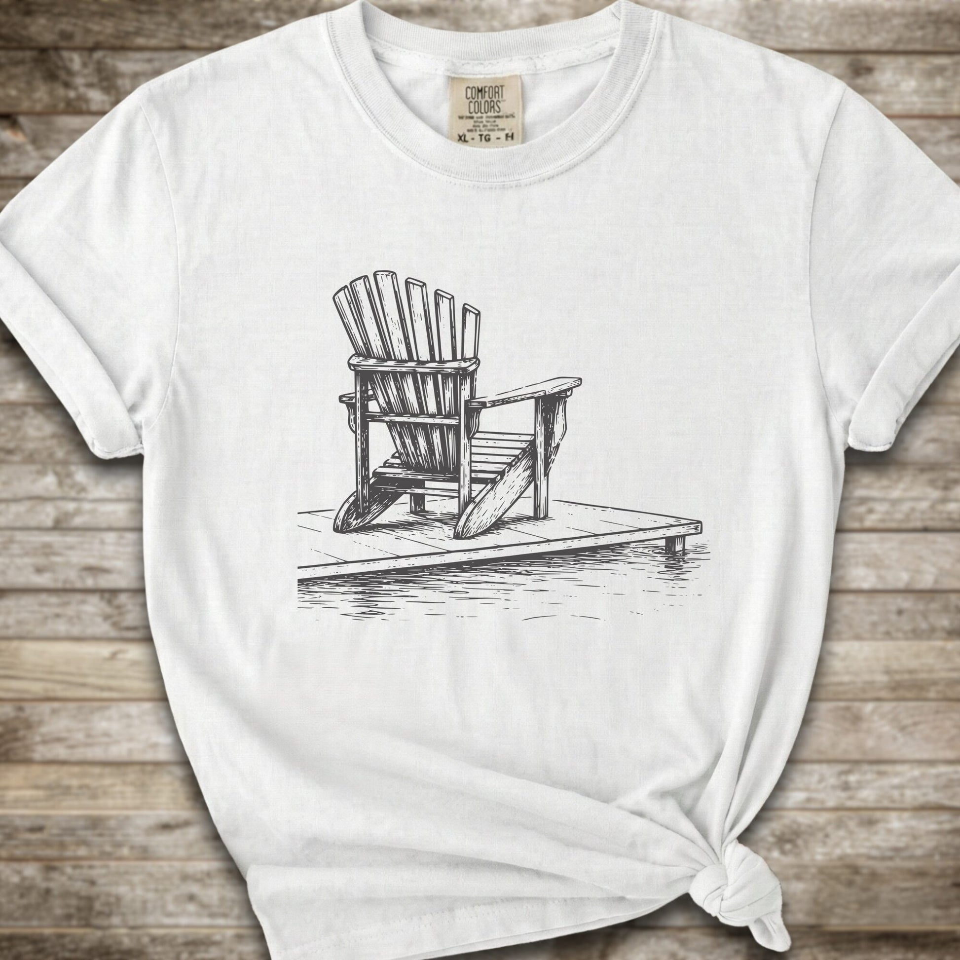 A white short-sleeve t-shirt with a white line art graphic of a Muskoka / Adirondack chair on a dock, styled with a knot at the bottom hem and laid flat on a rustic wood 