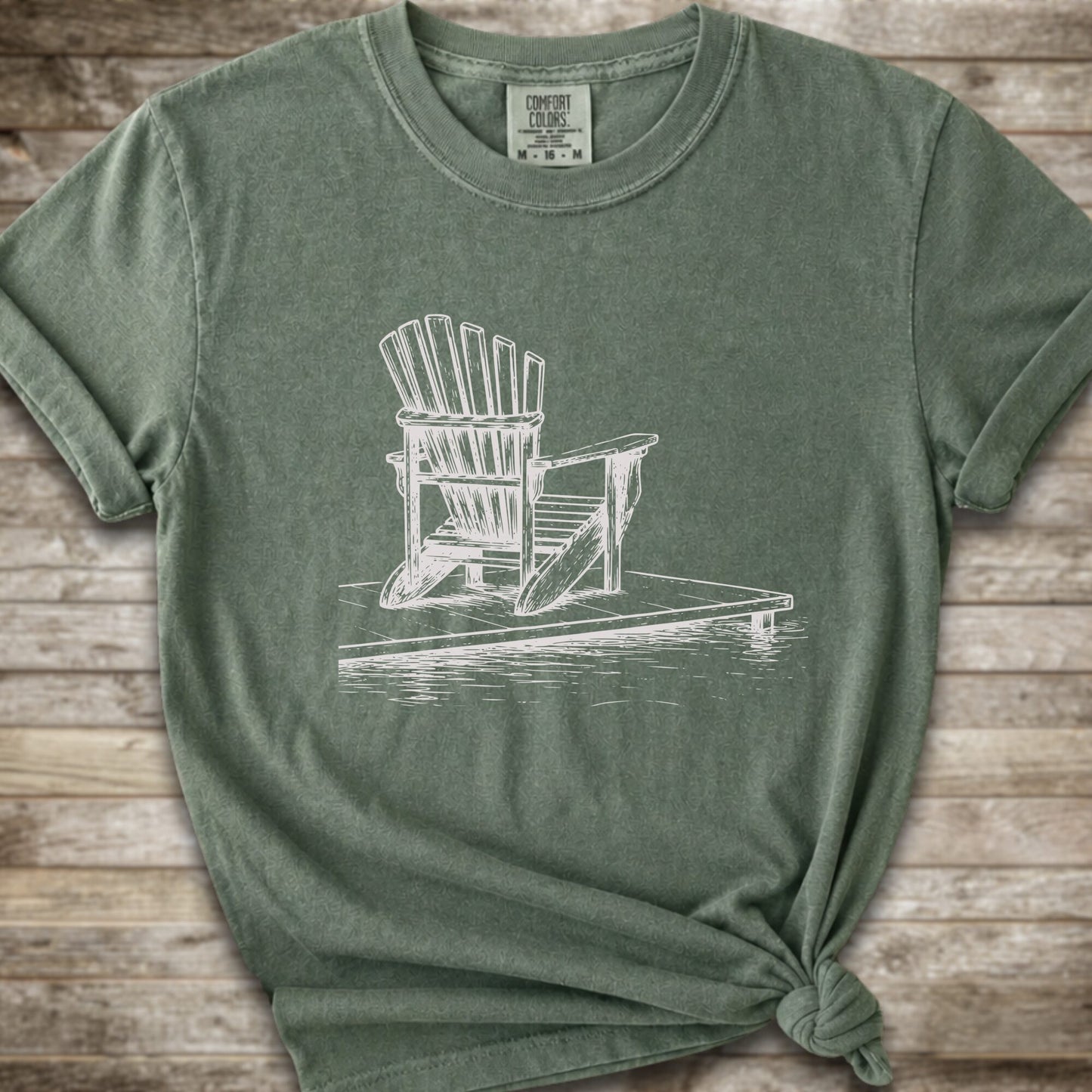 A moss green short-sleeve t-shirt with a white line art graphic of a Muskoka / Adirondack chair on a dock, styled with a knot at the bottom hem and laid flat on a rustic wood 