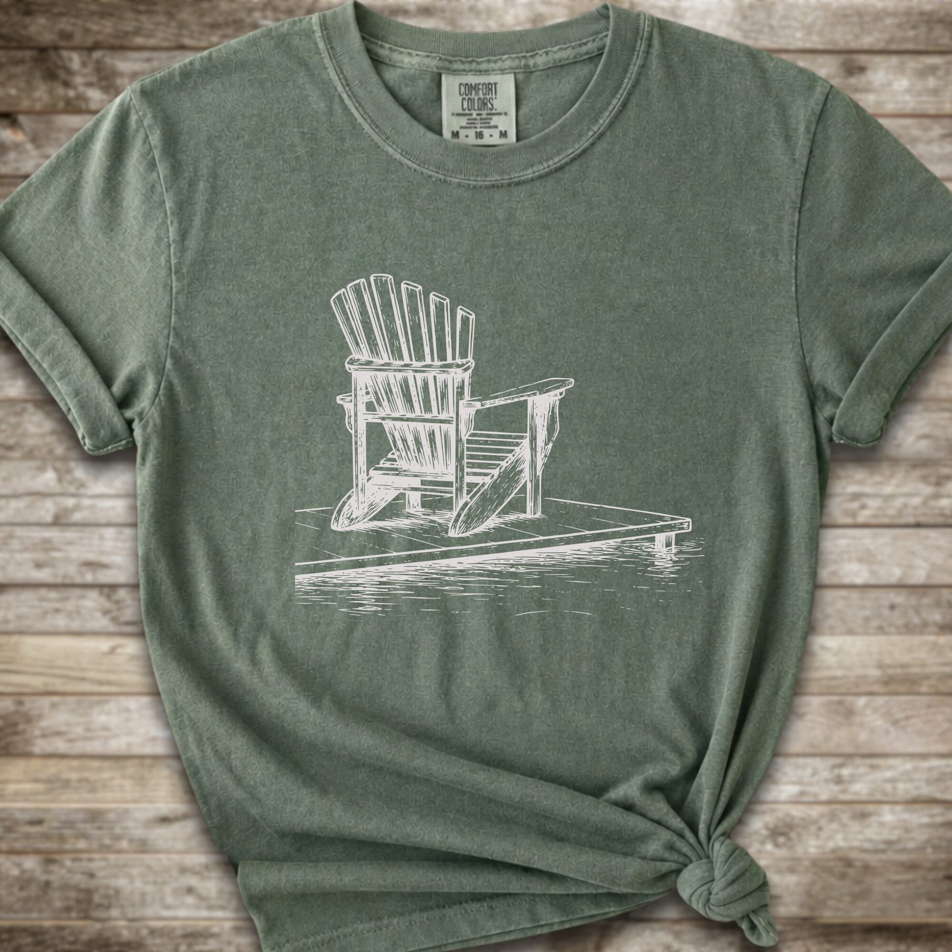 A moss green short-sleeve t-shirt with a white line art graphic of a Muskoka / Adirondack chair on a dock, styled with a knot at the bottom hem and laid flat on a rustic wood 