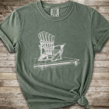 A moss green short-sleeve t-shirt with a white line art graphic of a Muskoka / Adirondack chair on a dock, styled with a knot at the bottom hem and laid flat on a rustic wood 