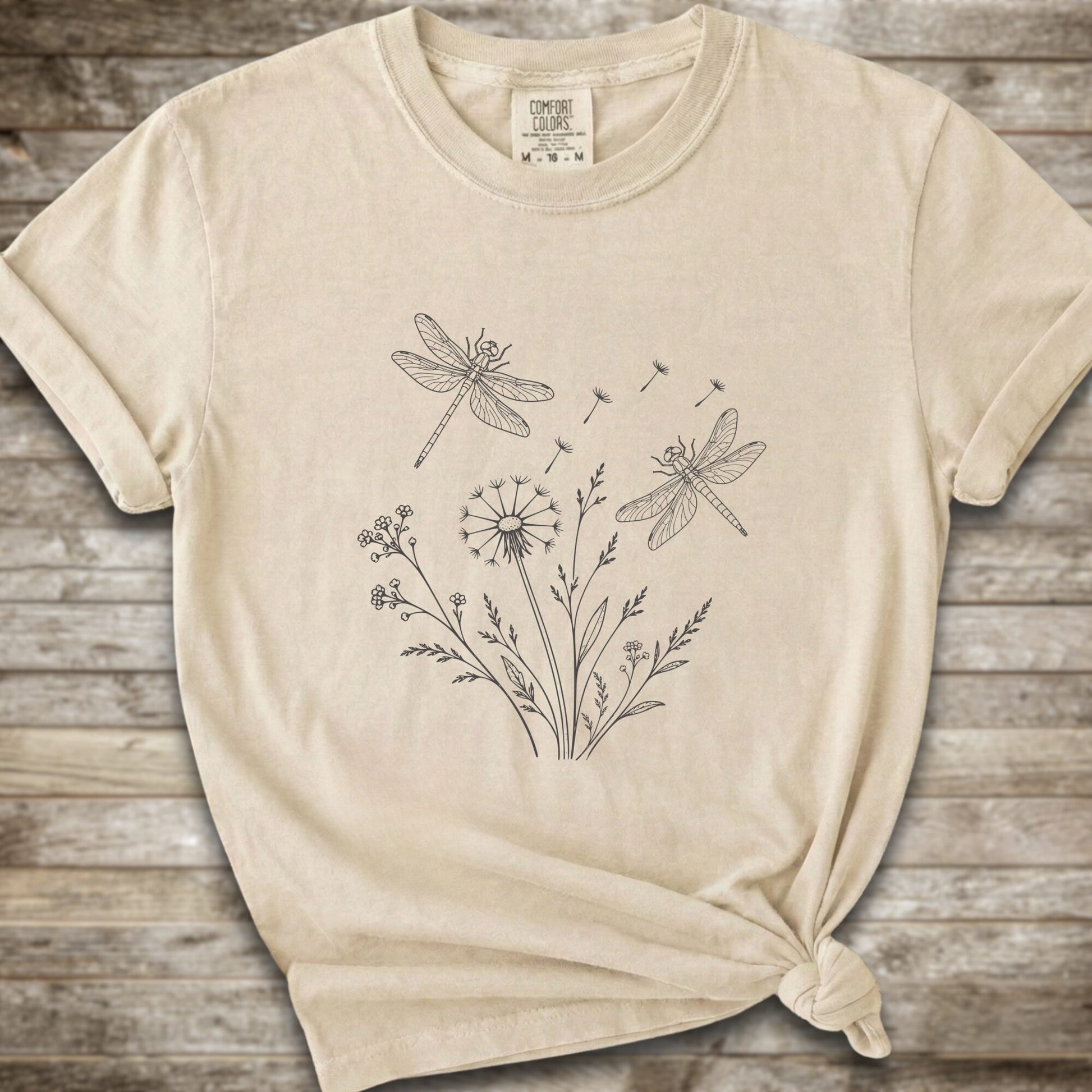 Ivory t-shirt with a white line art graphic of dragonflies, dandelions, and wildflowers, styled on a rustic wooden background.
