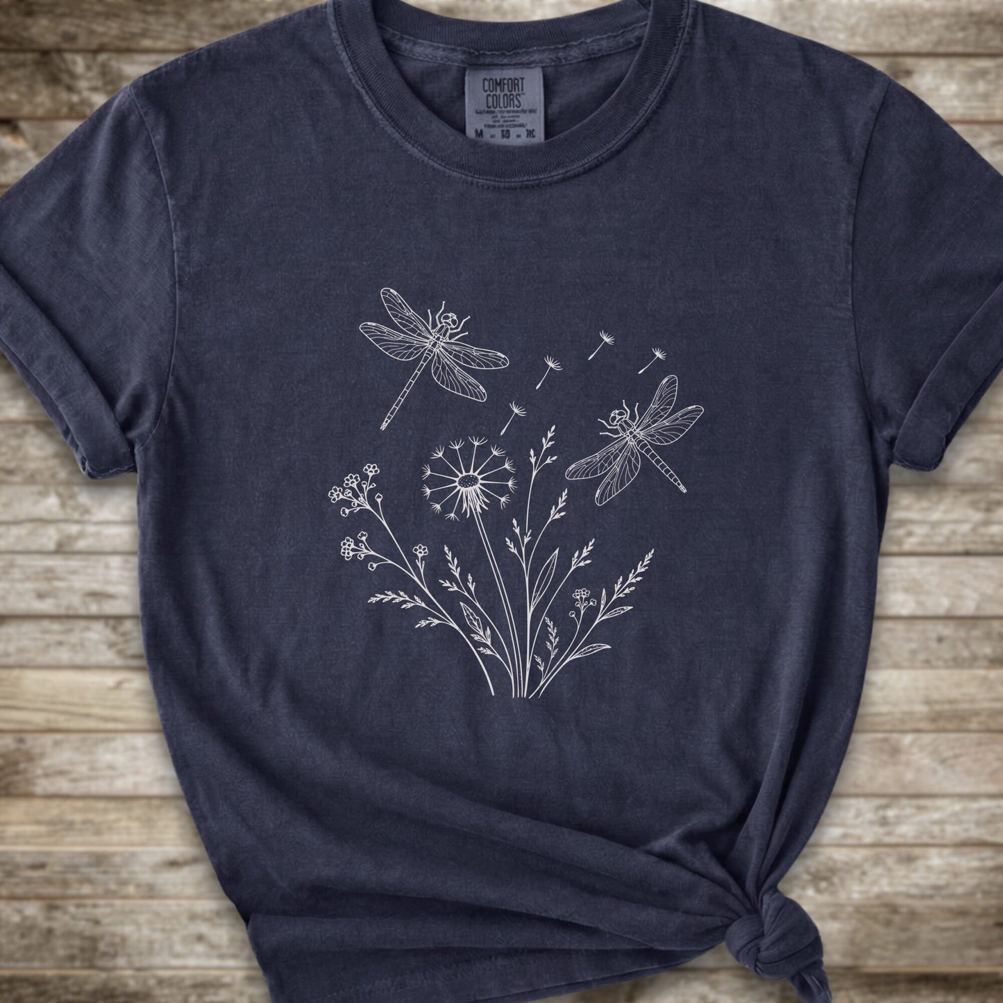 A dark navy blue t-shirt with a white line art graphic of dragonflies, dandelions, and wildflowers, styled on a rustic wooden background.