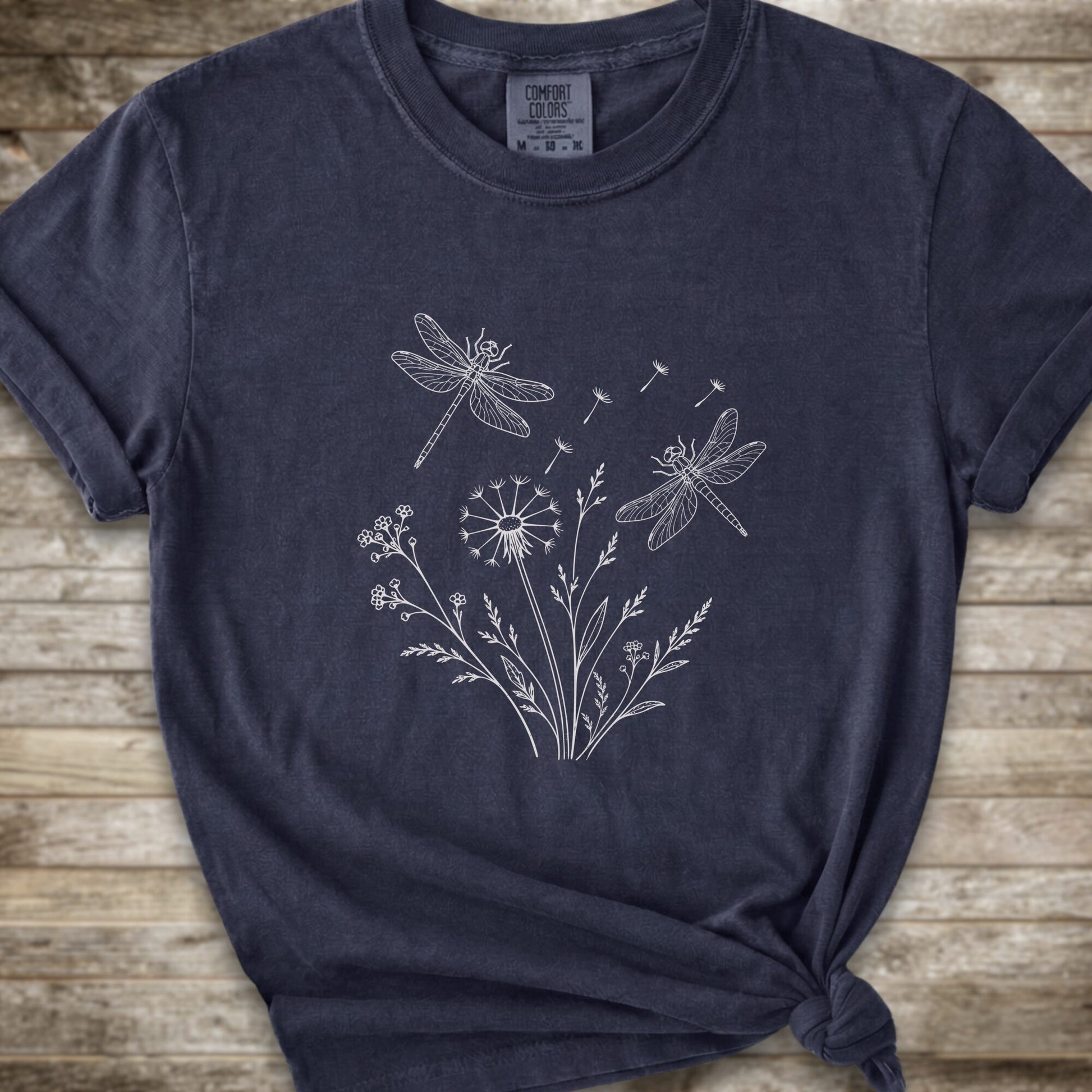 A dark navy blue t-shirt with a white line art graphic of dragonflies, dandelions, and wildflowers, styled on a rustic wooden background.