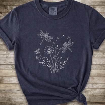 A dark navy blue t-shirt with a white line art graphic of dragonflies, dandelions, and wildflowers, styled on a rustic wooden background.