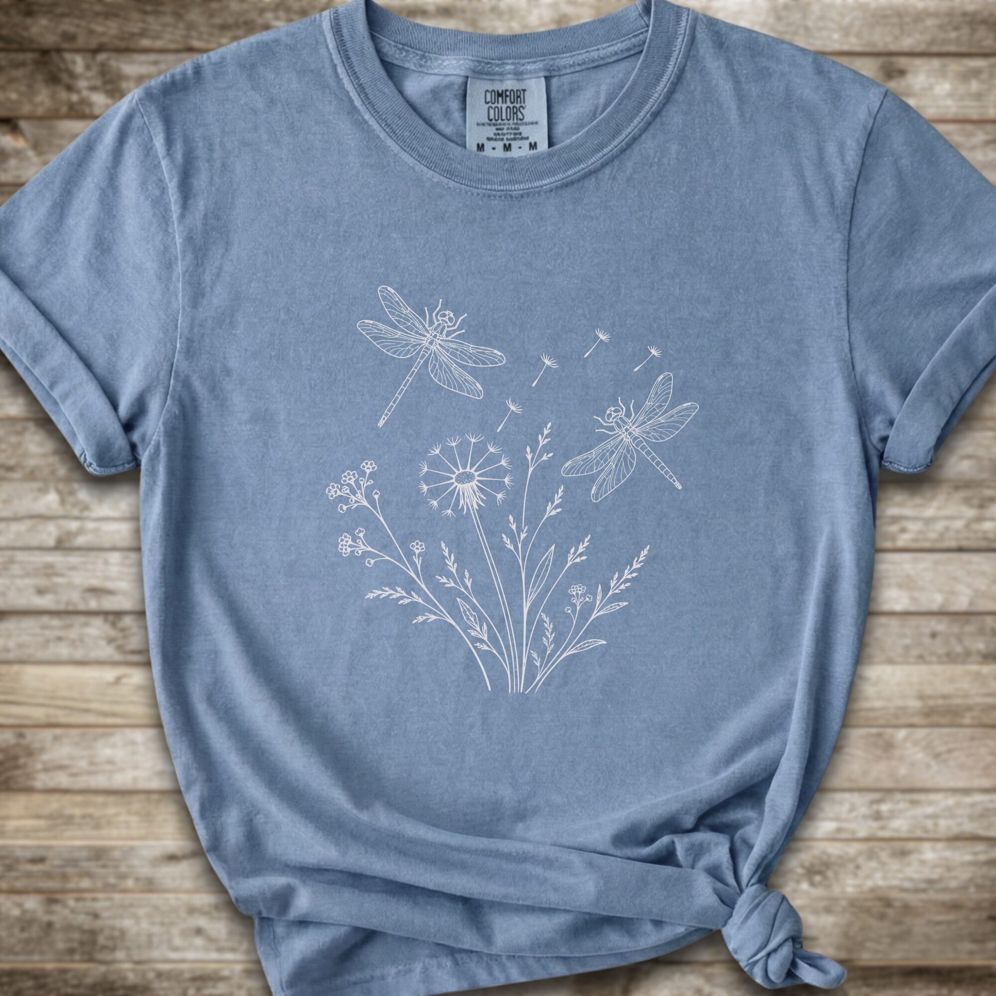 Light blue t-shirt with a white line art graphic of dragonflies, dandelions, and wildflowers, styled on a rustic wooden background.