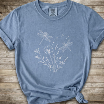 Light blue t-shirt with a white line art graphic of dragonflies, dandelions, and wildflowers, styled on a rustic wooden background.