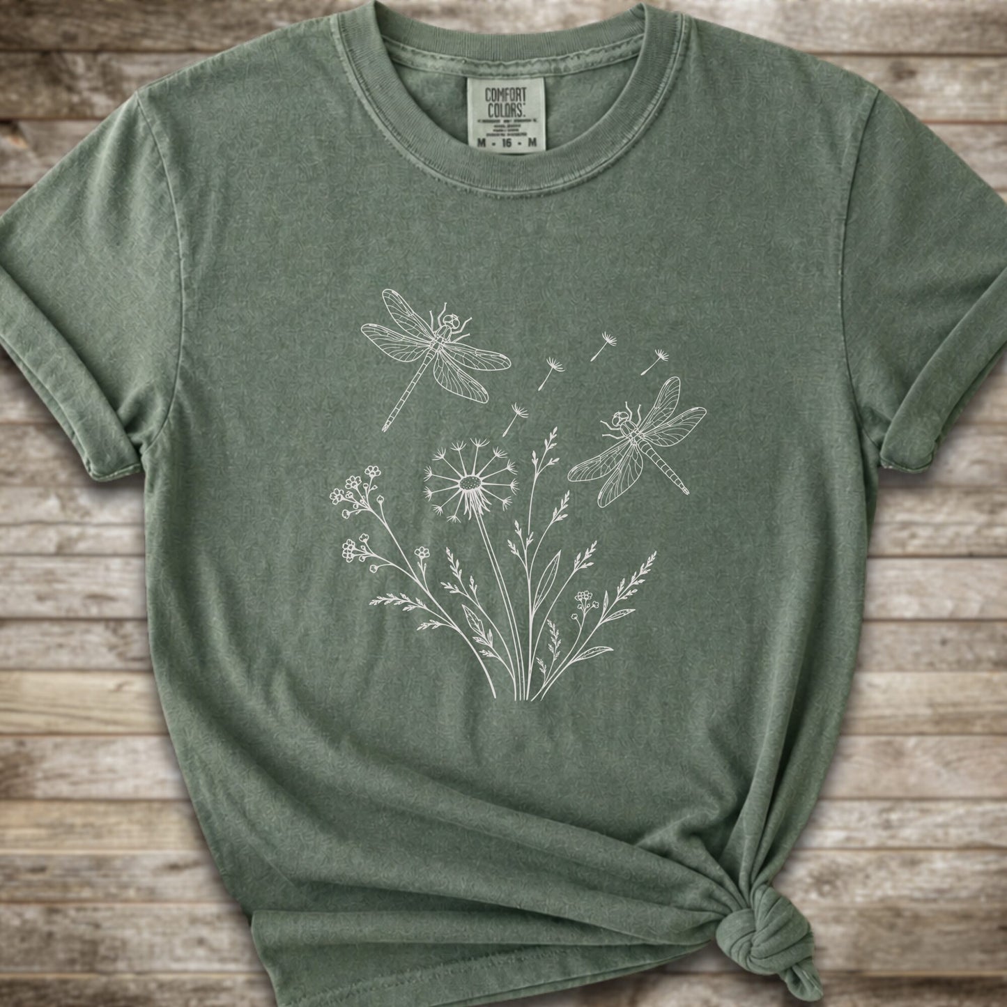 Moss t-shirt with a white line art graphic of dragonflies, dandelions, and wildflowers, styled on a rustic wooden background.