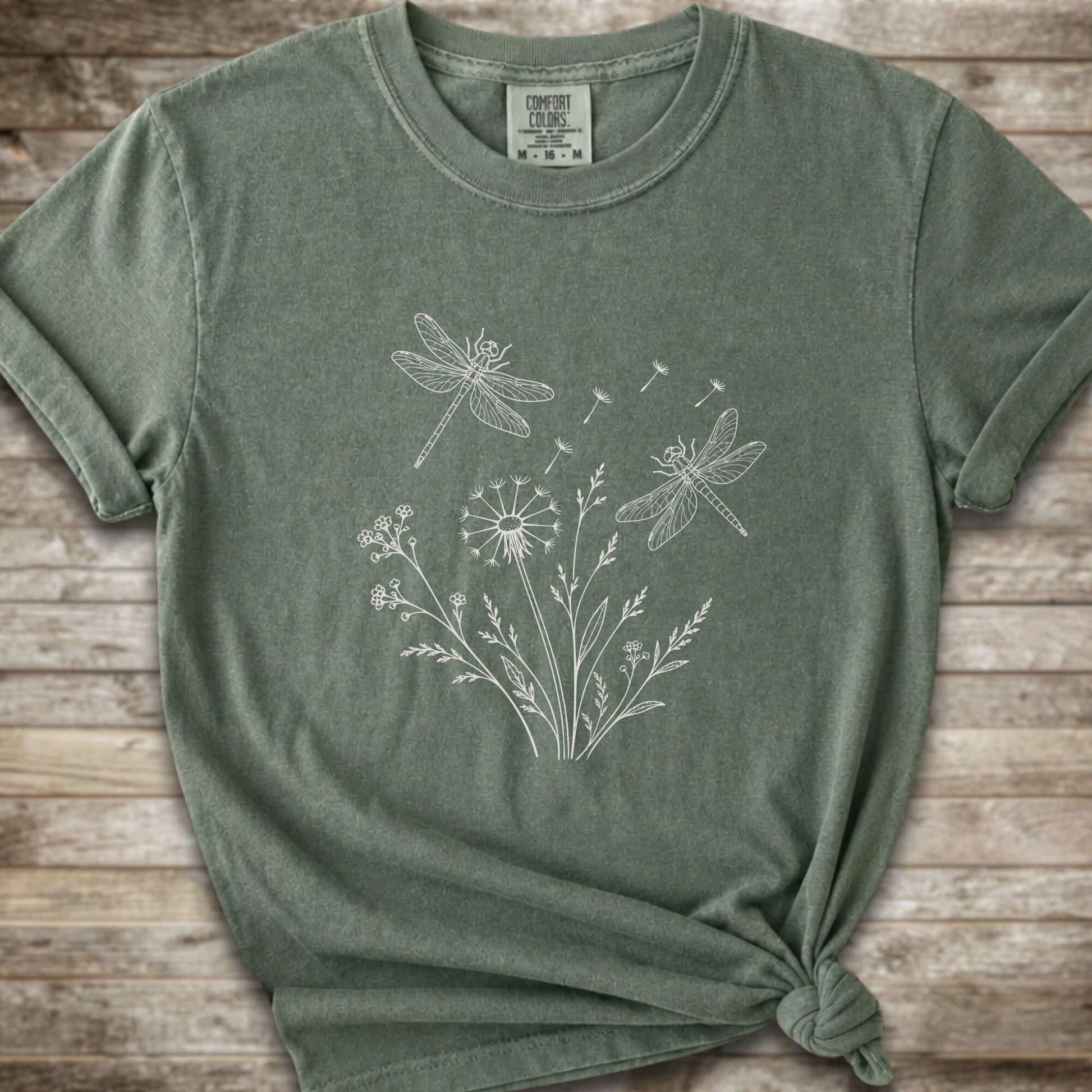 Moss t-shirt with a white line art graphic of dragonflies, dandelions, and wildflowers, styled on a rustic wooden background.