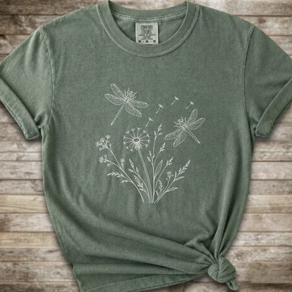 Moss t-shirt with a white line art graphic of dragonflies, dandelions, and wildflowers, styled on a rustic wooden background.
