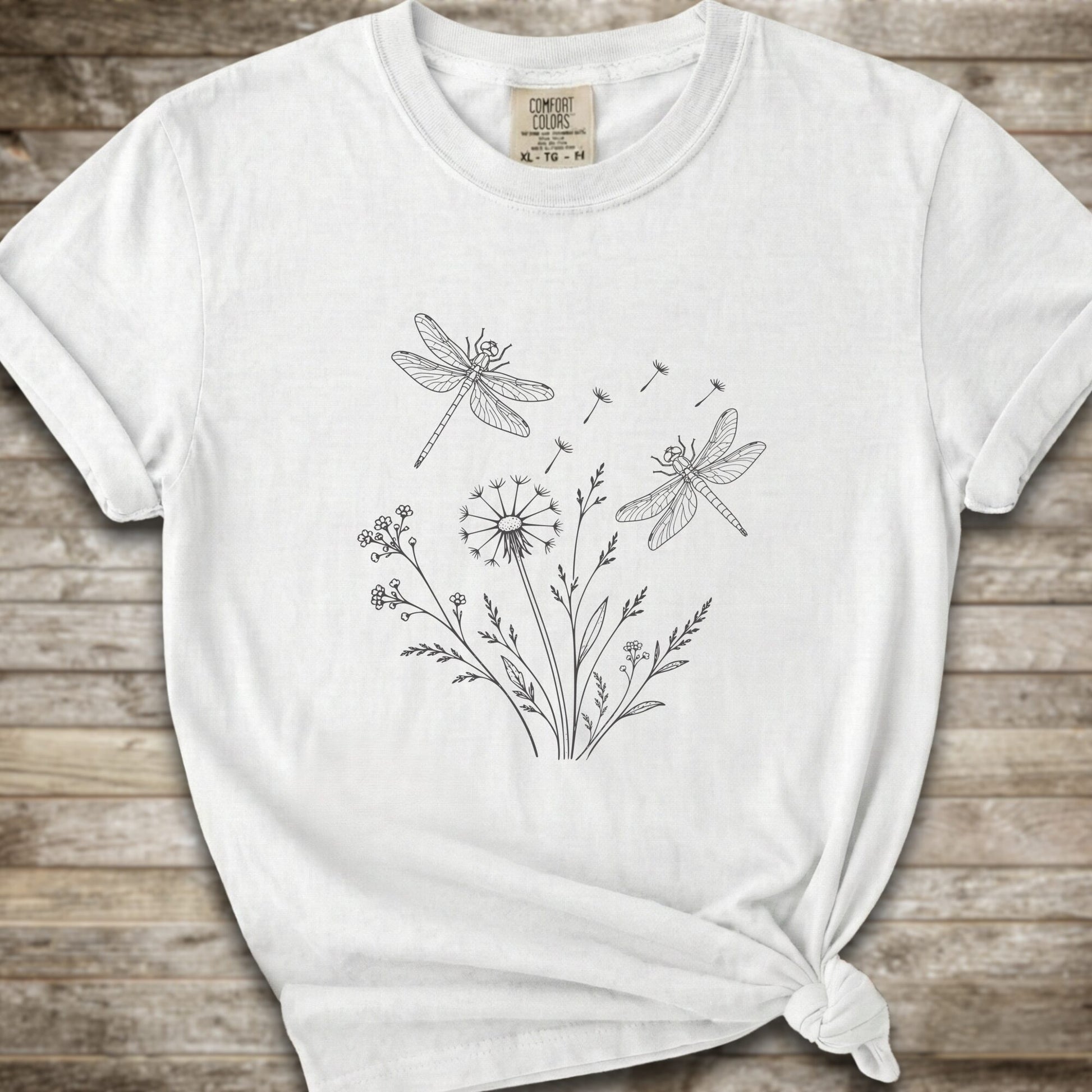 White t-shirt with a white line art graphic of dragonflies, dandelions, and wildflowers, styled on a rustic wooden background.