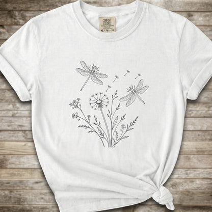 White t-shirt with a white line art graphic of dragonflies, dandelions, and wildflowers, styled on a rustic wooden background.