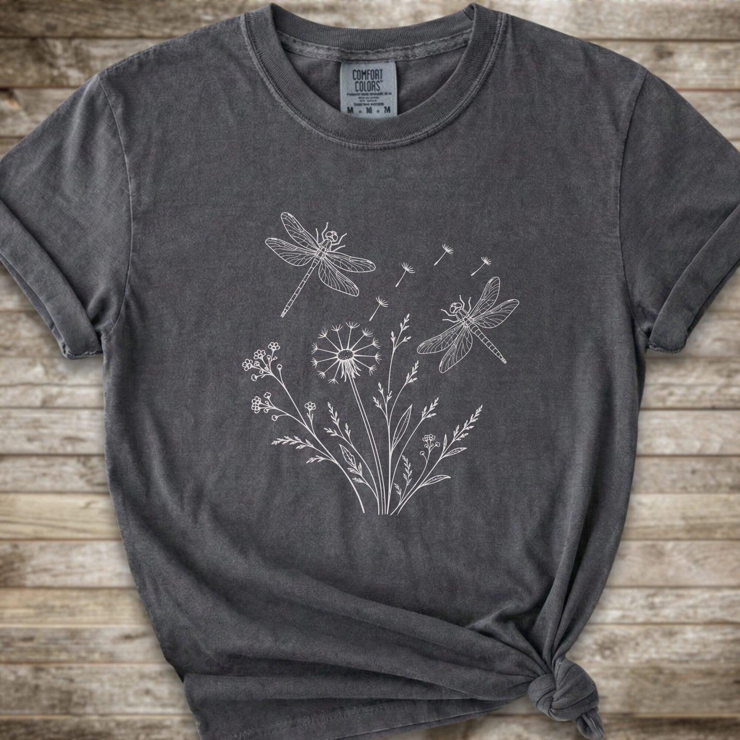 Dark Grey t-shirt with a white line art graphic of dragonflies, dandelions, and wildflowers, styled on a rustic wooden background.