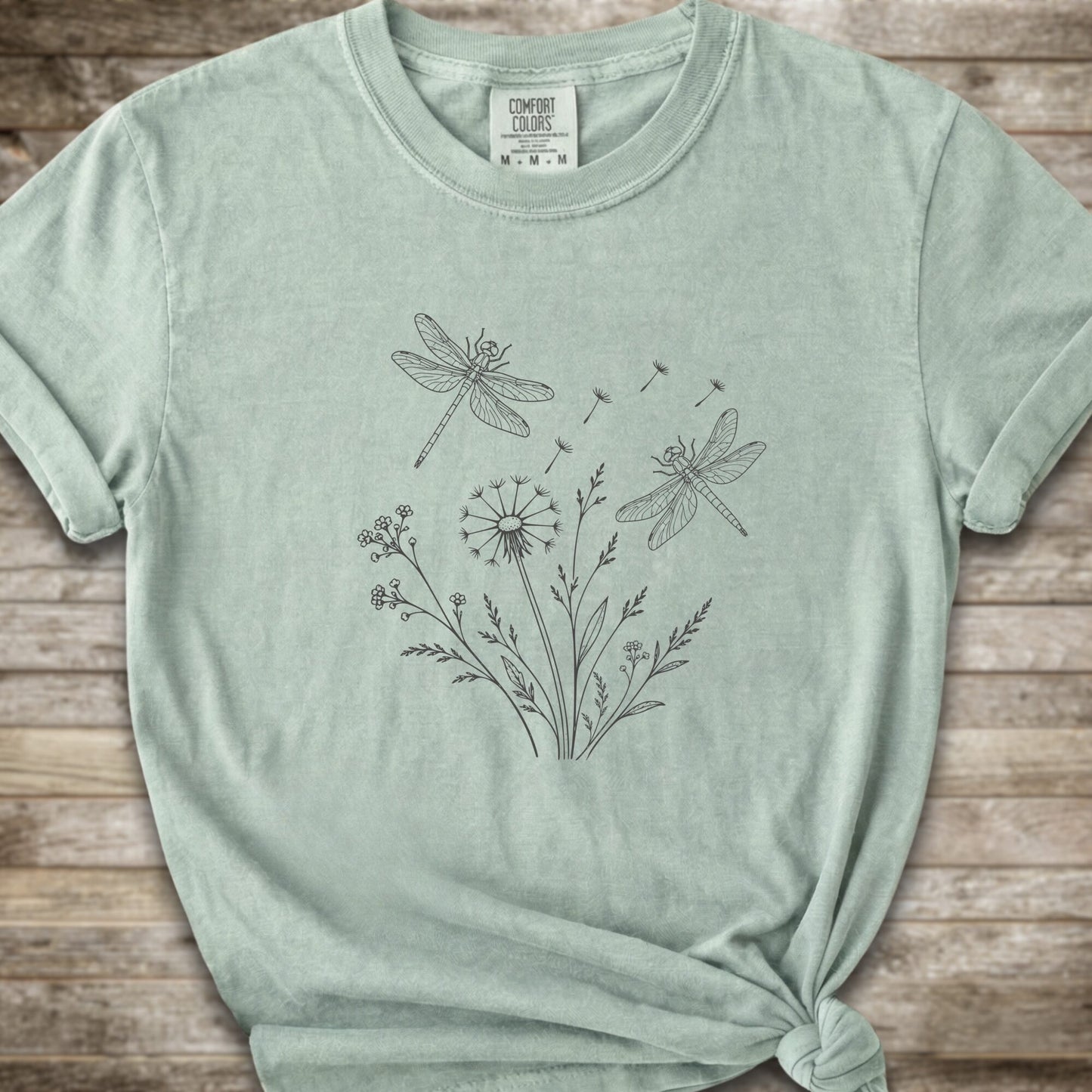 Light Green t-shirt with a white line art graphic of dragonflies, dandelions, and wildflowers, styled on a rustic wooden background.
