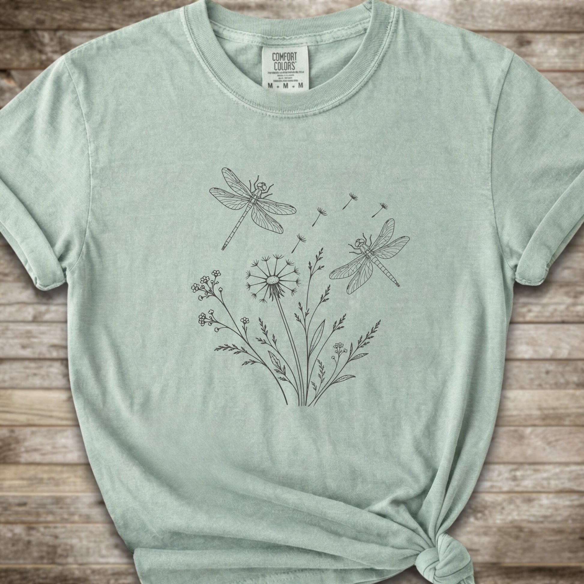 Light Green t-shirt with a white line art graphic of dragonflies, dandelions, and wildflowers, styled on a rustic wooden background.