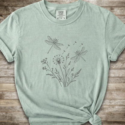 Light Green t-shirt with a white line art graphic of dragonflies, dandelions, and wildflowers, styled on a rustic wooden background.