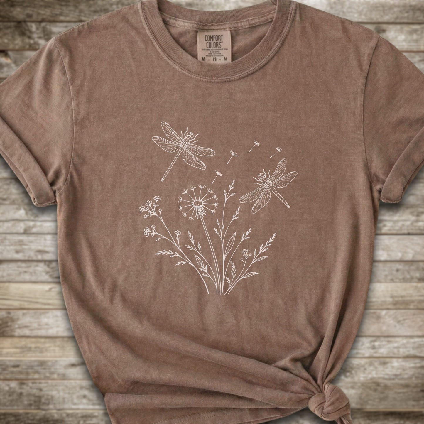 Brown t-shirt with a white line art graphic of dragonflies, dandelions, and wildflowers, styled on a rustic wooden background.
