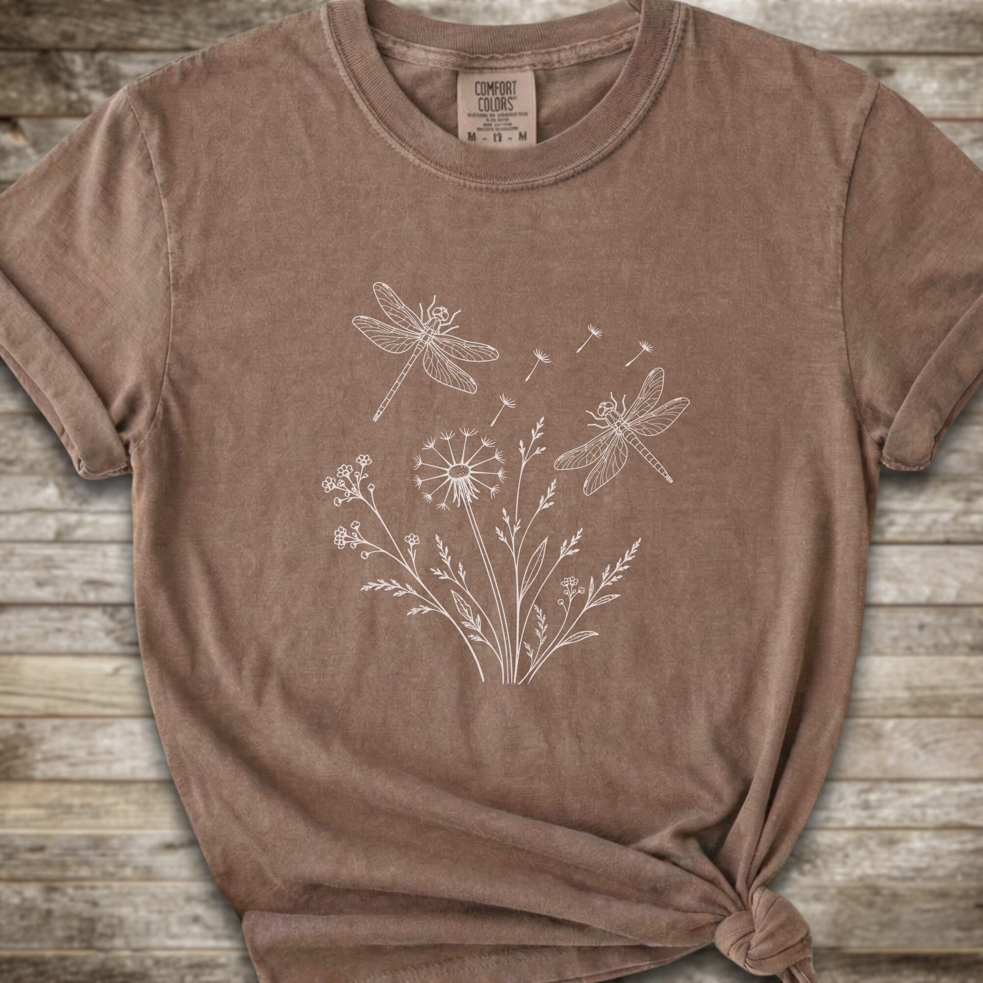 Brown t-shirt with a white line art graphic of dragonflies, dandelions, and wildflowers, styled on a rustic wooden background.