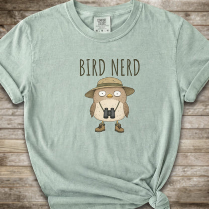 A light sage green t-shirt with a graphic of a cute cartoon bird wearing a safari hat and binoculars, with the text 'BIRD NERD' printed above it. The t-shirt is laid flat on a rustic wooden background and is styled with a knot at the bottom right hem.