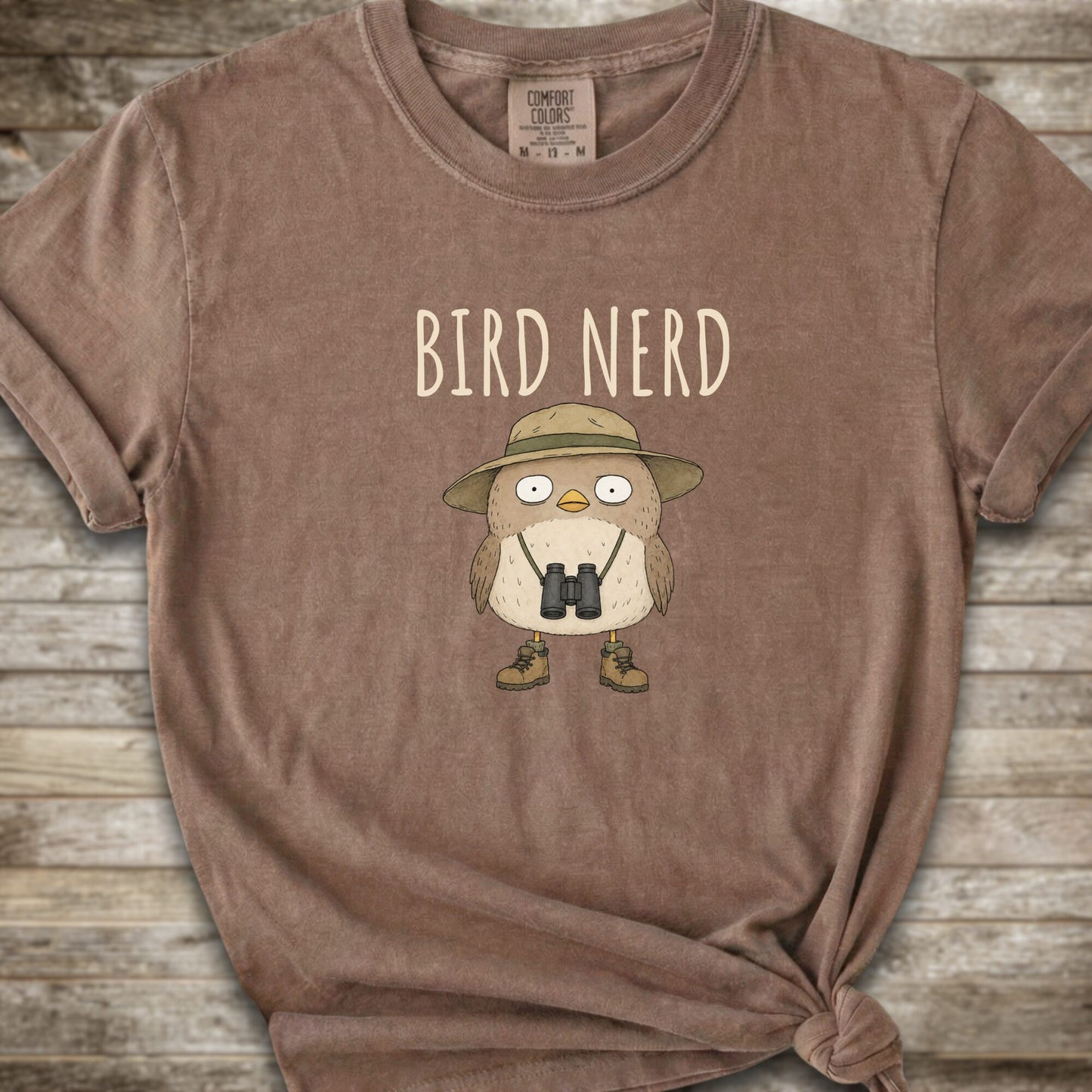 Brown t-shirt with a graphic of a cute cartoon bird wearing a safari hat and binoculars, with the text 'BIRD NERD' printed above it. The t-shirt is laid flat on a rustic wooden background and is styled with a knot at the bottom right hem.