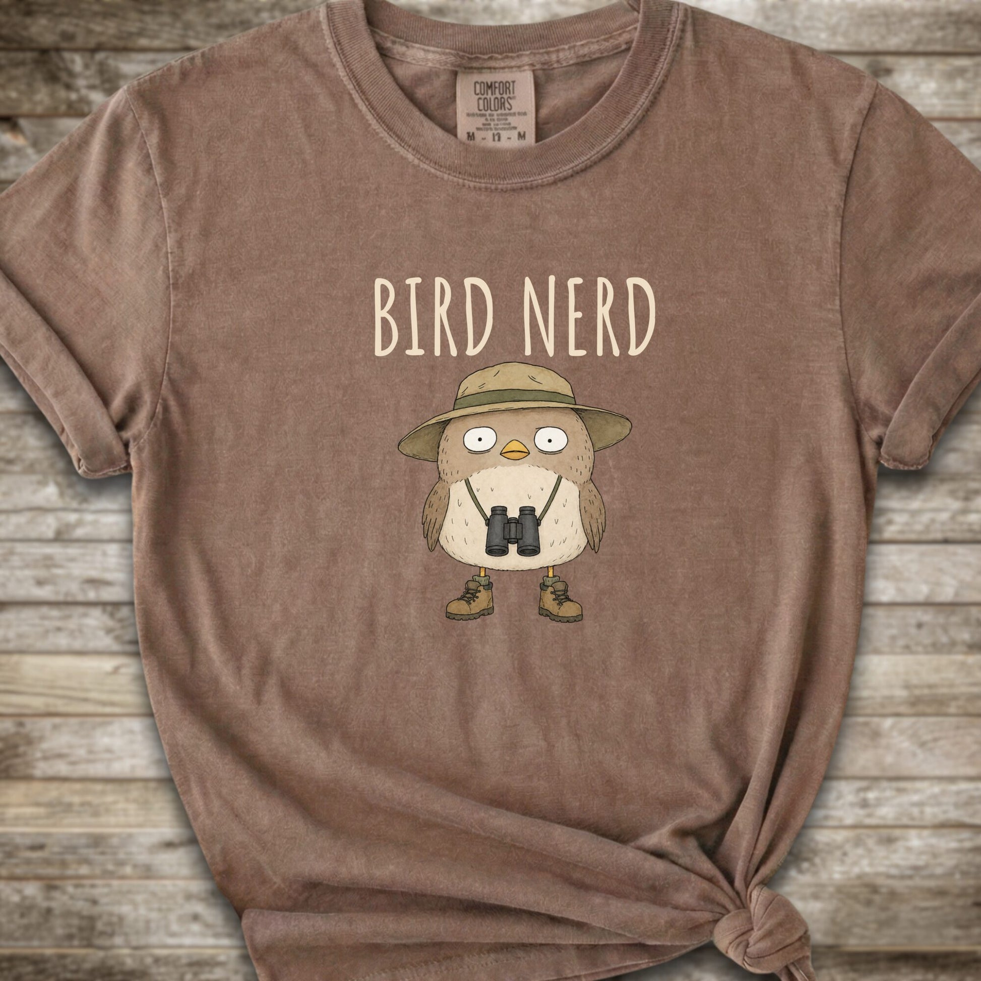Brown t-shirt with a graphic of a cute cartoon bird wearing a safari hat and binoculars, with the text 'BIRD NERD' printed above it. The t-shirt is laid flat on a rustic wooden background and is styled with a knot at the bottom right hem.