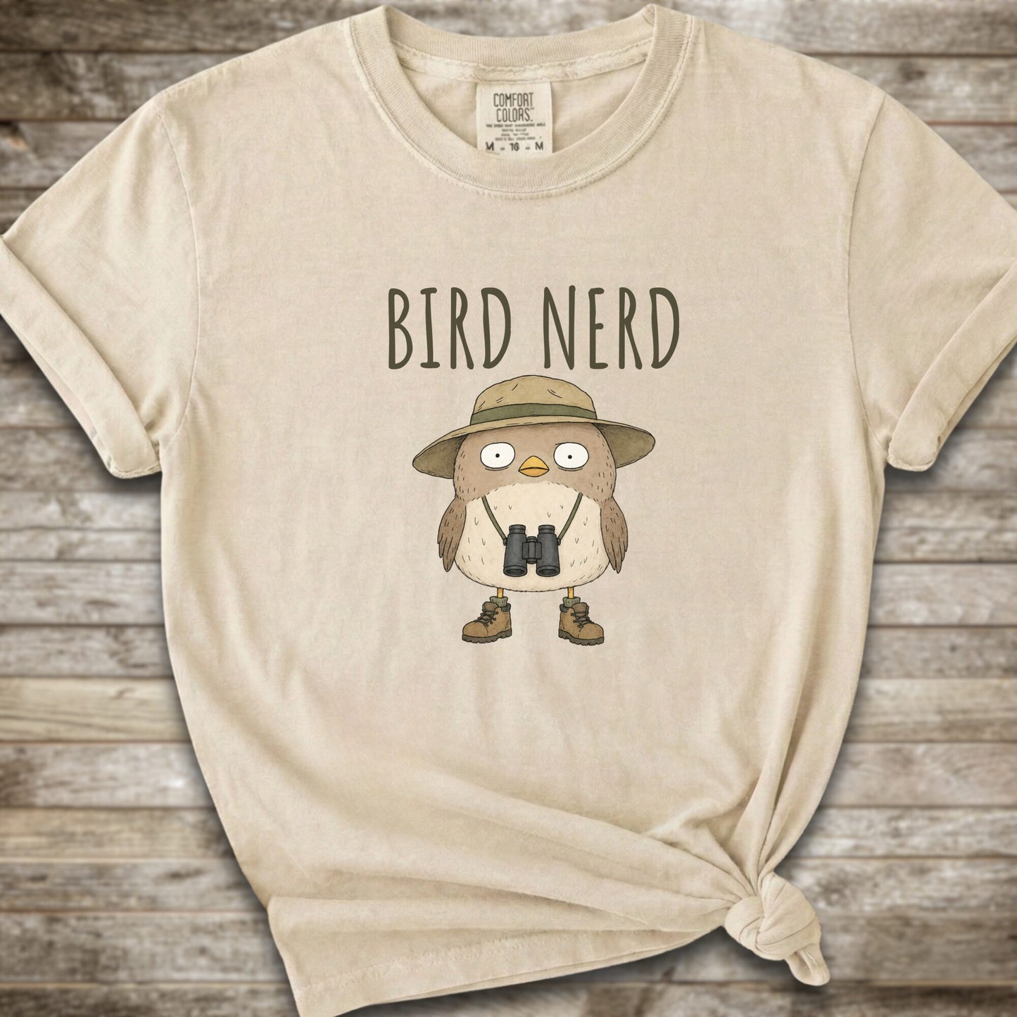 Ivory t-shirt with a graphic of a cute cartoon bird wearing a safari hat and binoculars, with the text 'BIRD NERD' printed above it. The t-shirt is laid flat on a rustic wooden background and is styled with a knot at the bottom right hem.