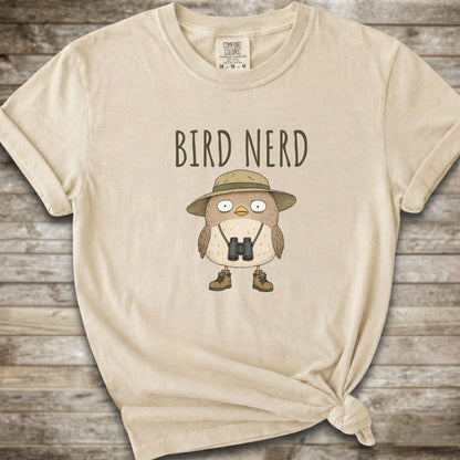 Ivory t-shirt with a graphic of a cute cartoon bird wearing a safari hat and binoculars, with the text 'BIRD NERD' printed above it. The t-shirt is laid flat on a rustic wooden background and is styled with a knot at the bottom right hem.
