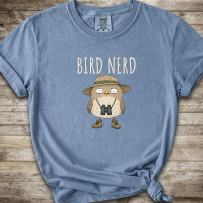 Blue t-shirt with a graphic of a cute cartoon bird wearing a safari hat and binoculars, with the text 'BIRD NERD' printed above it. The t-shirt is laid flat on a rustic wooden background and is styled with a knot at the bottom right hem.