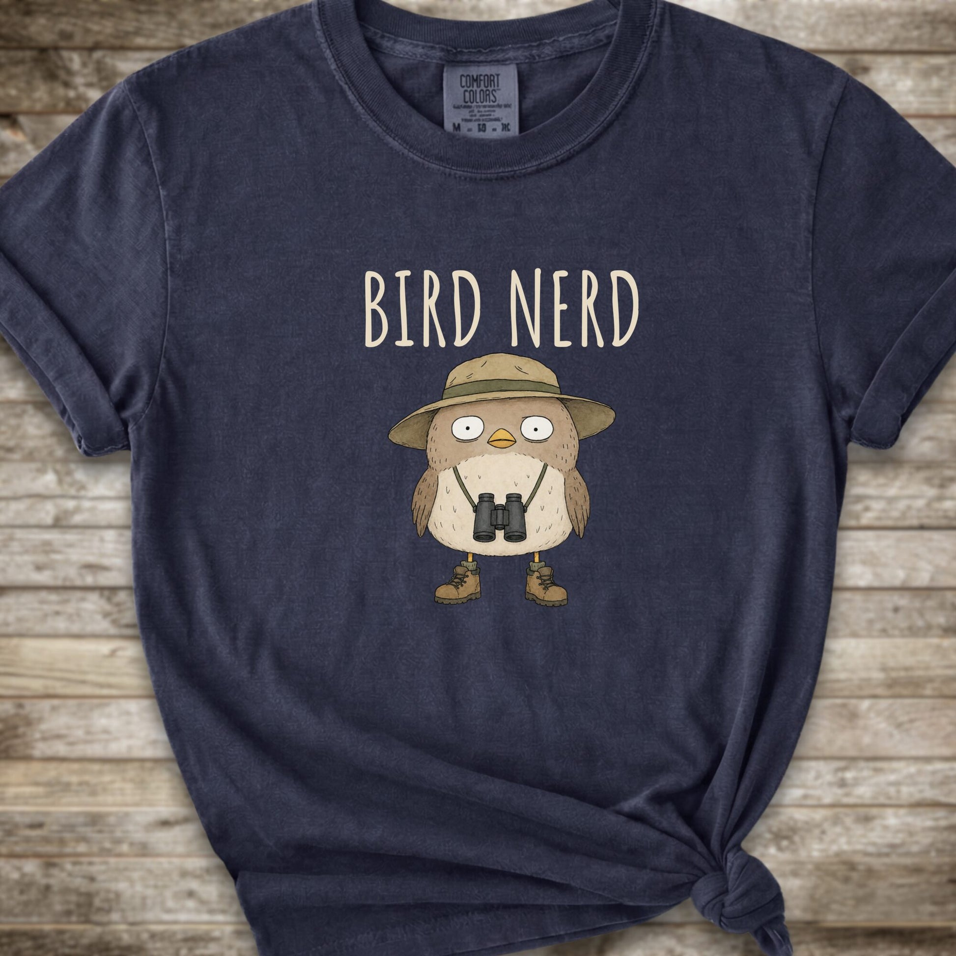 Navy blue t-shirt with a graphic of a cute cartoon bird wearing a safari hat and binoculars, with the text 'BIRD NERD' printed above it. The t-shirt is laid flat on a rustic wooden background and is styled with a knot at the bottom right hem.