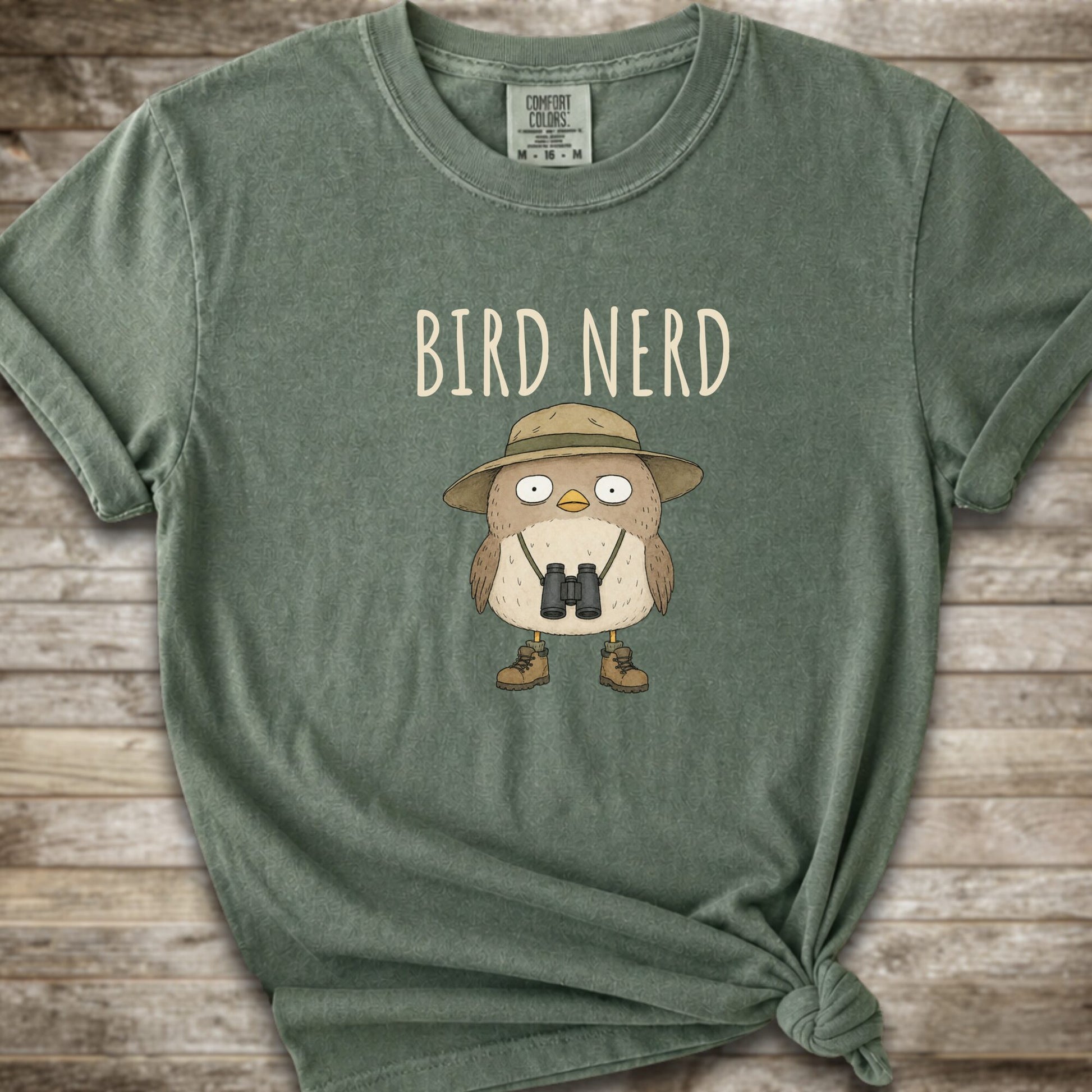 Moss green t-shirt with a graphic of a cute cartoon bird wearing a safari hat and binoculars, with the text 'BIRD NERD' printed above it. The t-shirt is laid flat on a rustic wooden background and is styled with a knot at the bottom right hem.