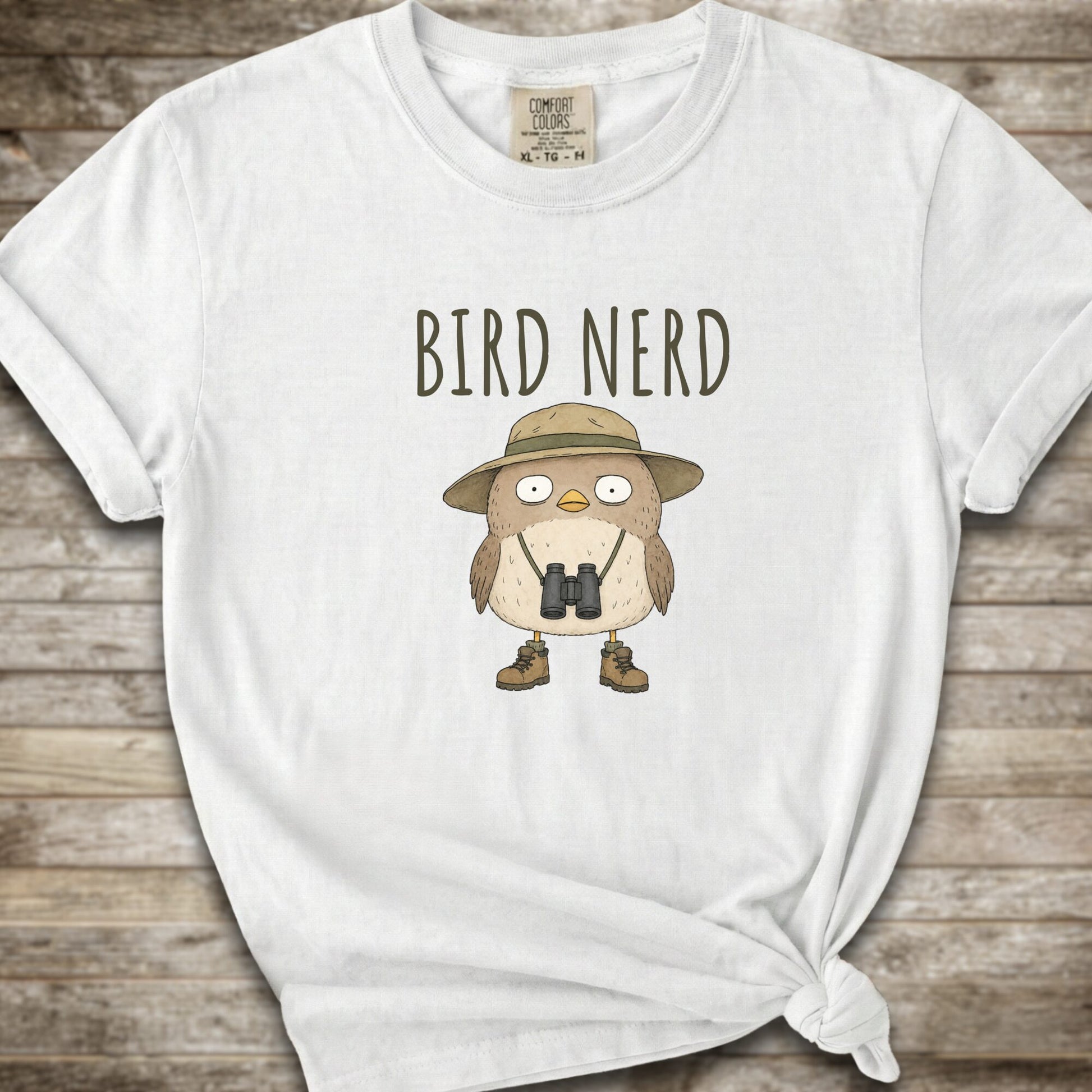 Whte t-shirt with a graphic of a cute cartoon bird wearing a safari hat and binoculars, with the text 'BIRD NERD' printed above it. The t-shirt is laid flat on a rustic wooden background and is styled with a knot at the bottom right hem.