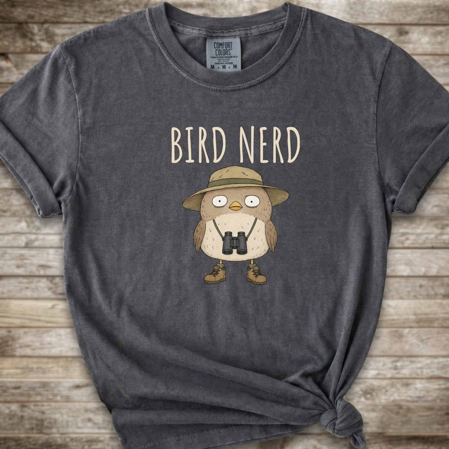 Dark Grey t-shirt with a graphic of a cute cartoon bird wearing a safari hat and binoculars, with the text 'BIRD NERD' printed above it. The t-shirt is laid flat on a rustic wooden background and is styled with a knot at the bottom right hem.