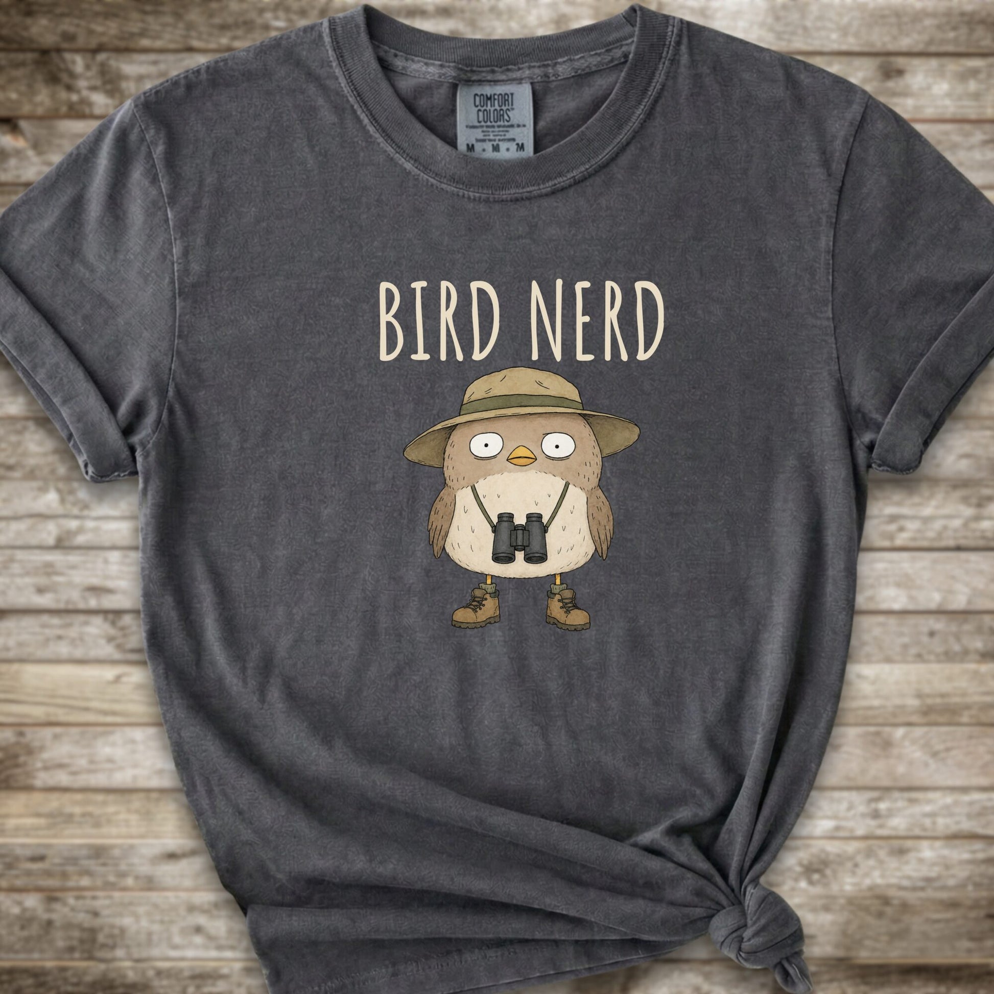 Dark Grey t-shirt with a graphic of a cute cartoon bird wearing a safari hat and binoculars, with the text 'BIRD NERD' printed above it. The t-shirt is laid flat on a rustic wooden background and is styled with a knot at the bottom right hem.