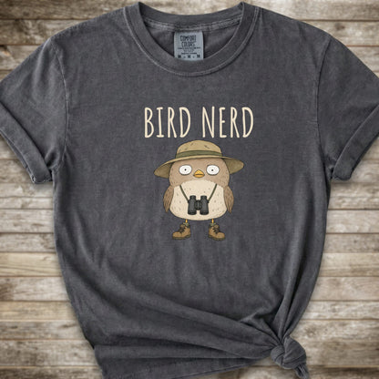 Dark Grey t-shirt with a graphic of a cute cartoon bird wearing a safari hat and binoculars, with the text 'BIRD NERD' printed above it. The t-shirt is laid flat on a rustic wooden background and is styled with a knot at the bottom right hem.