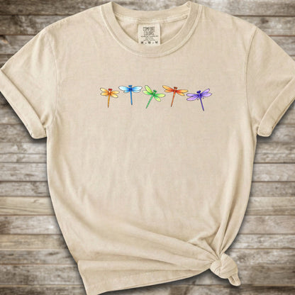 Ivory short-sleeve t-shirt with a graphic of five colorful dragonflies across the chest, styled with a knot at the bottom hem and laid flat on a rustic wooden background.