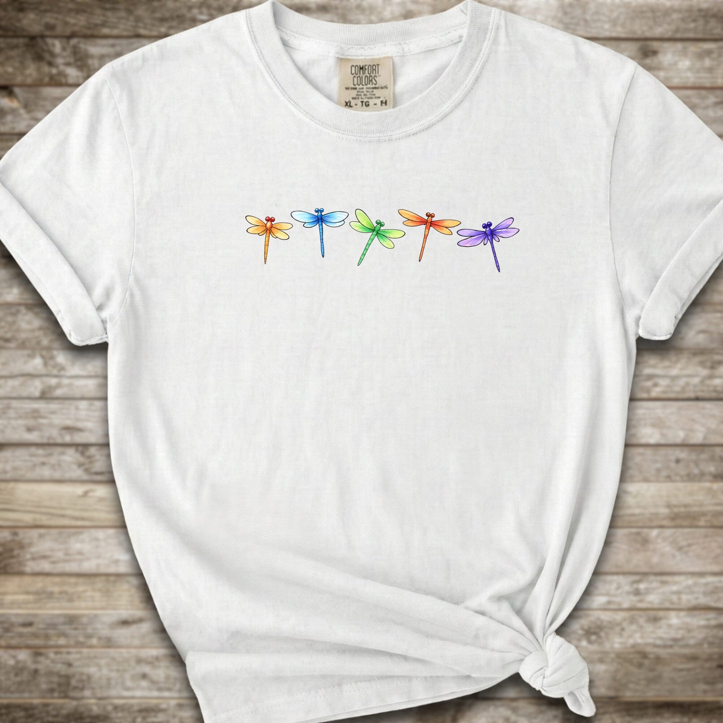 A white short-sleeve t-shirt with a graphic of five colorful dragonflies across the chest, styled with a knot at the bottom hem and laid flat on a rustic wooden background.