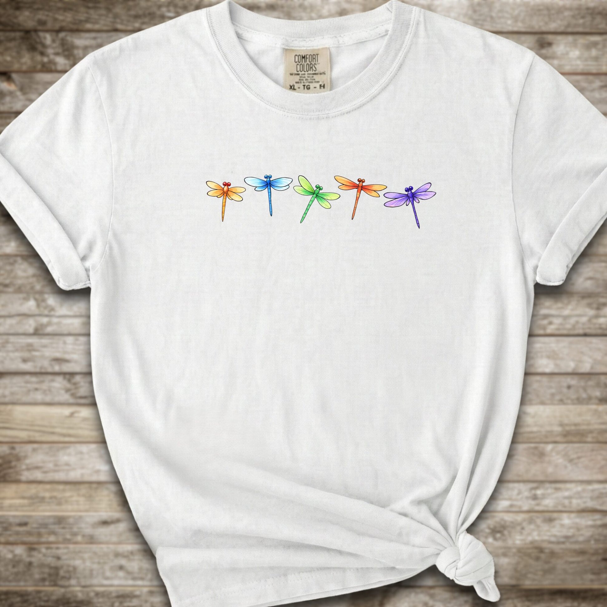 A white short-sleeve t-shirt with a graphic of five colorful dragonflies across the chest, styled with a knot at the bottom hem and laid flat on a rustic wooden background.