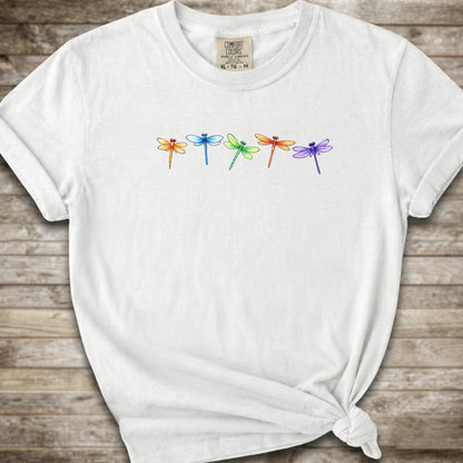 A white short-sleeve t-shirt with a graphic of five colorful dragonflies across the chest, styled with a knot at the bottom hem and laid flat on a rustic wooden background.