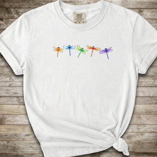 A white short-sleeve t-shirt with a graphic of five colorful dragonflies across the chest, styled with a knot at the bottom hem and laid flat on a rustic wooden background.