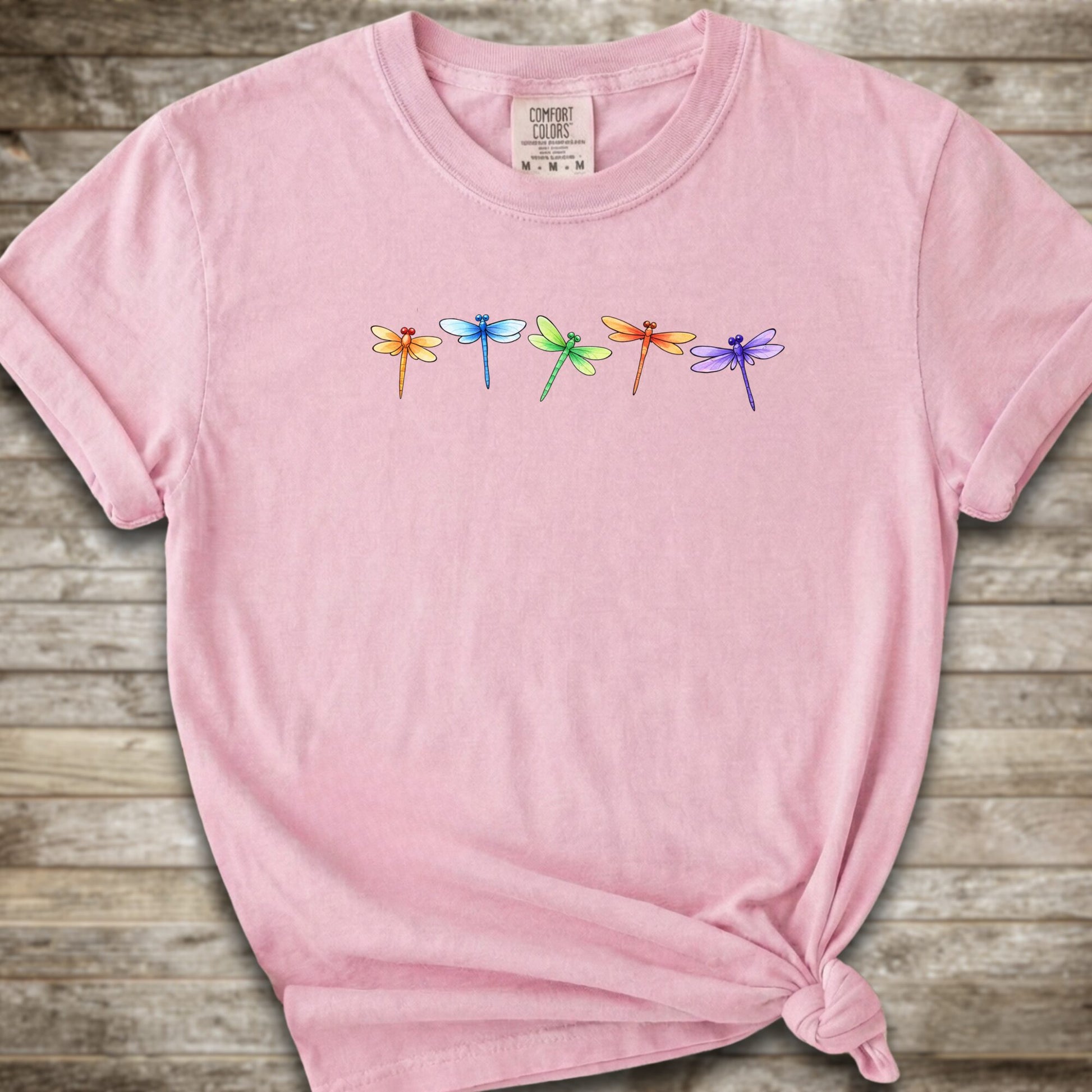Pink short-sleeve t-shirt with a graphic of five colorful dragonflies across the chest, styled with a knot at the bottom hem and laid flat on a rustic wooden background.