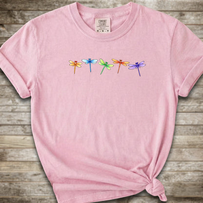 Pink short-sleeve t-shirt with a graphic of five colorful dragonflies across the chest, styled with a knot at the bottom hem and laid flat on a rustic wooden background.