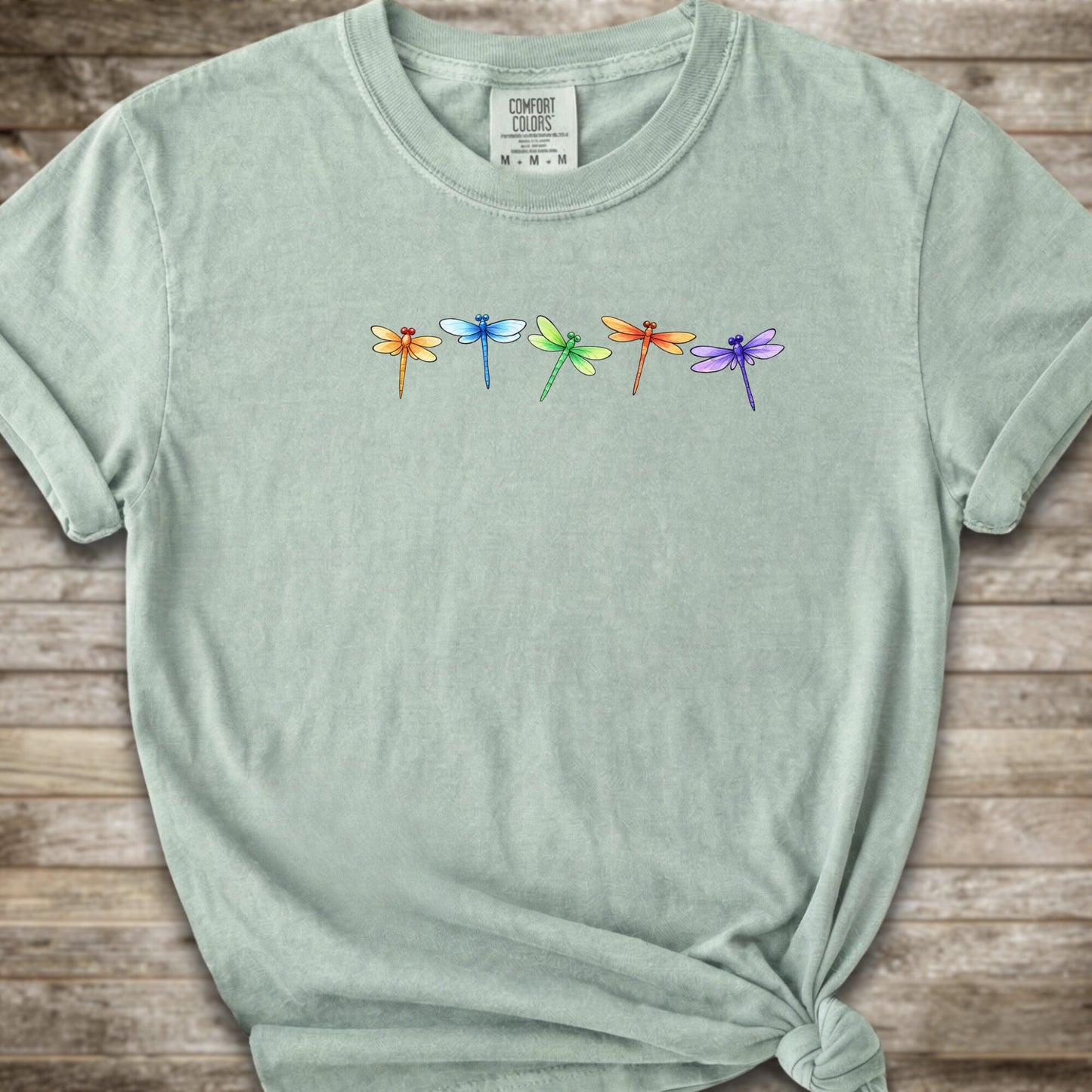 Green short-sleeve t-shirt with a graphic of five colorful dragonflies across the chest, styled with a knot at the bottom hem and laid flat on a rustic wooden background.