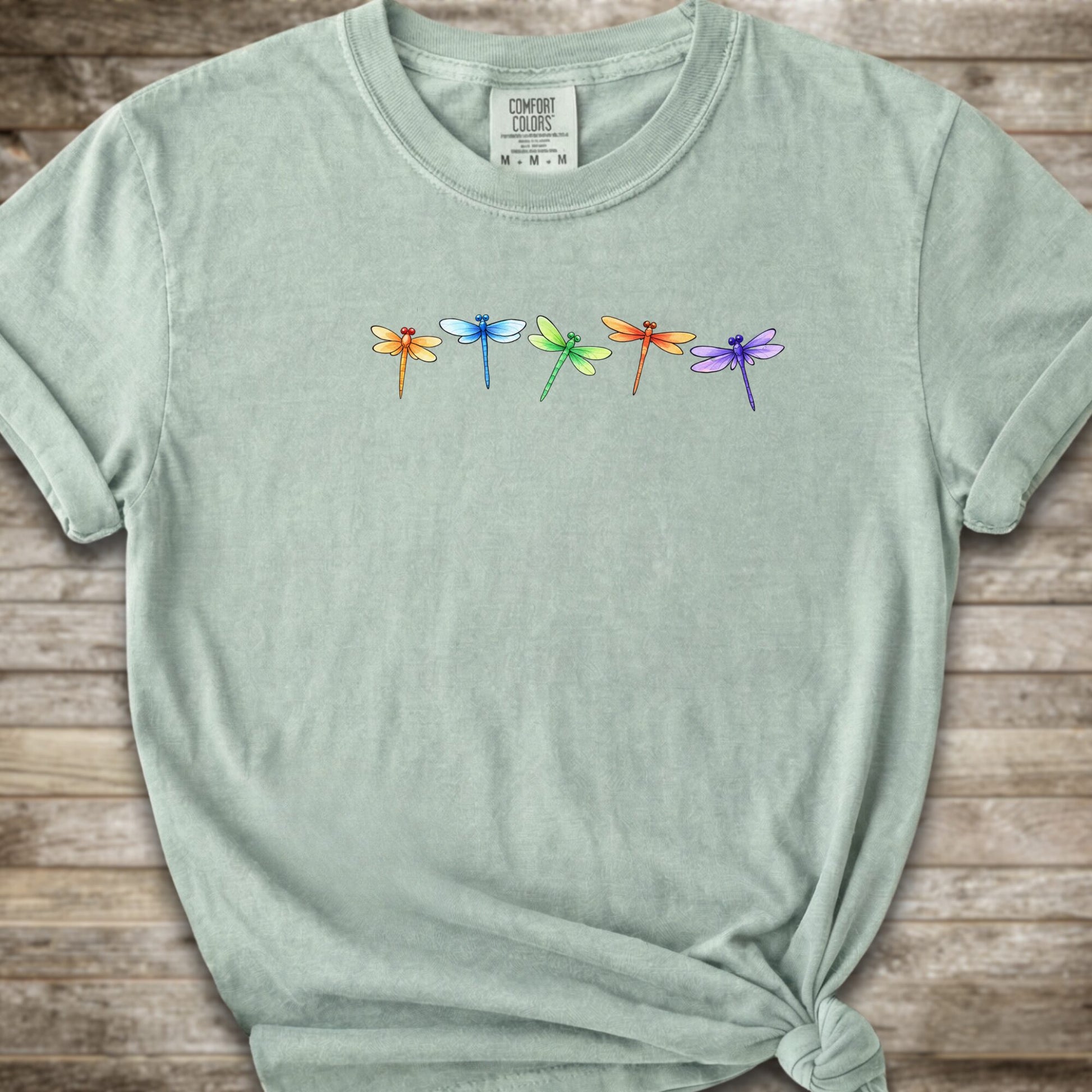 Green short-sleeve t-shirt with a graphic of five colorful dragonflies across the chest, styled with a knot at the bottom hem and laid flat on a rustic wooden background.