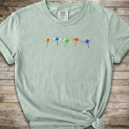 Green short-sleeve t-shirt with a graphic of five colorful dragonflies across the chest, styled with a knot at the bottom hem and laid flat on a rustic wooden background.