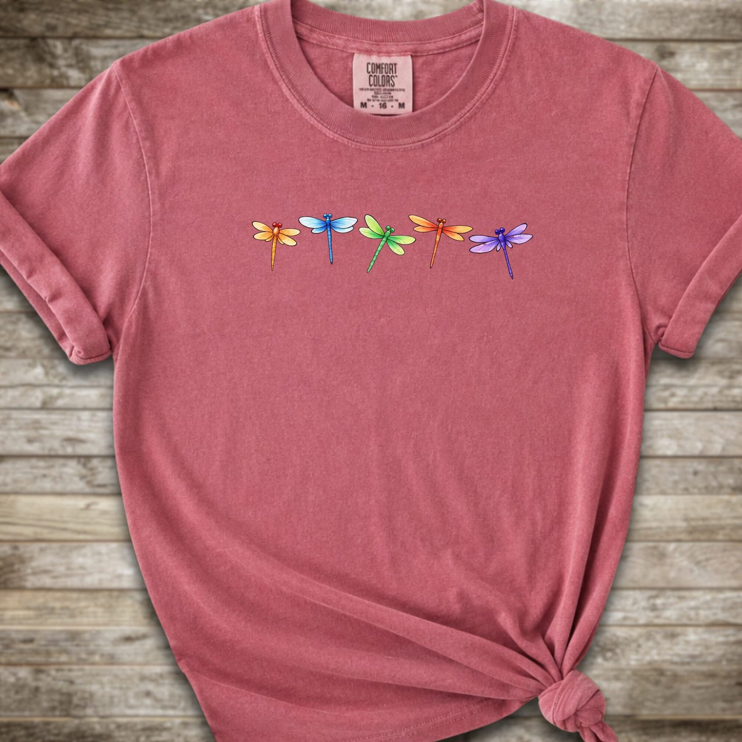 Dark Pink short-sleeve t-shirt with a graphic of five colorful dragonflies across the chest, styled with a knot at the bottom hem and laid flat on a rustic wooden background.