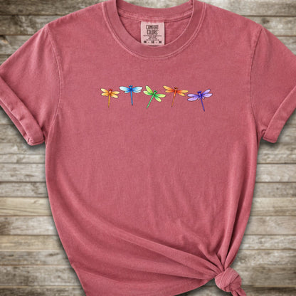 Dark Pink short-sleeve t-shirt with a graphic of five colorful dragonflies across the chest, styled with a knot at the bottom hem and laid flat on a rustic wooden background.