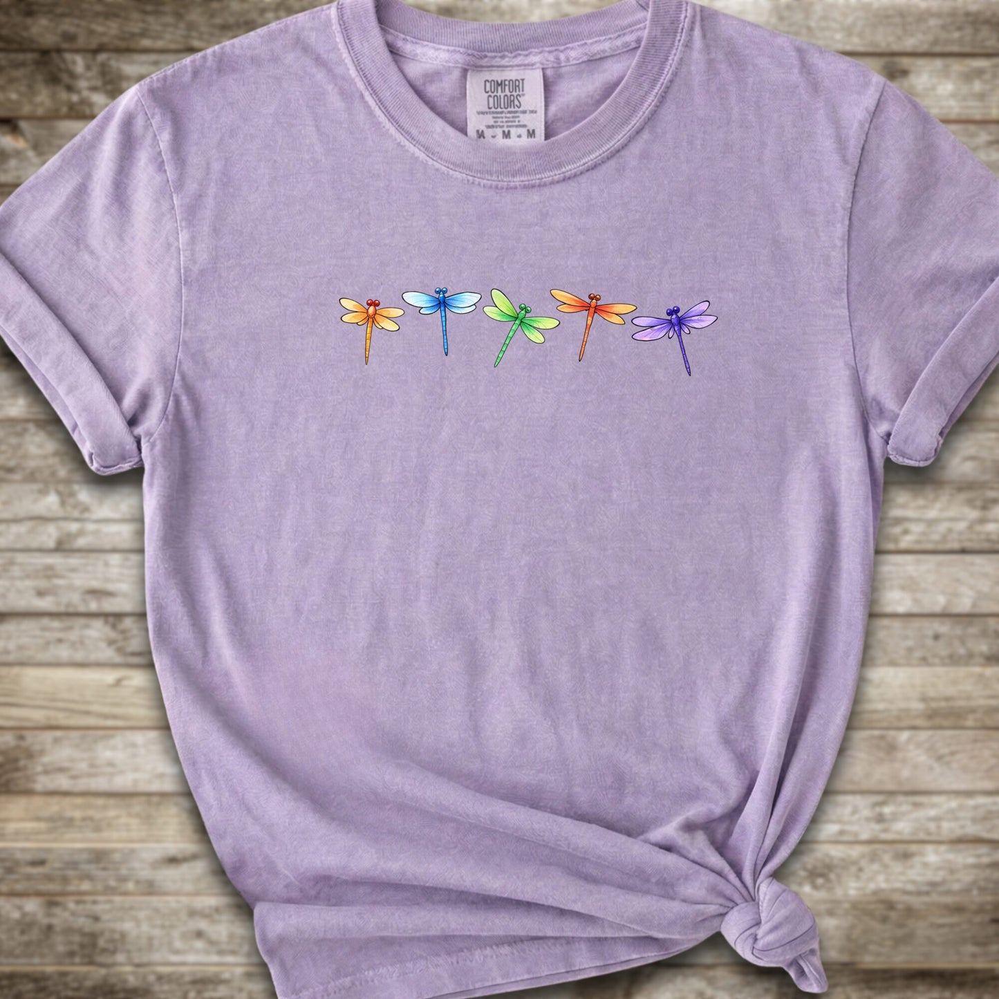 Purple short-sleeve t-shirt with a graphic of five colorful dragonflies across the chest, styled with a knot at the bottom hem and laid flat on a rustic wooden background.