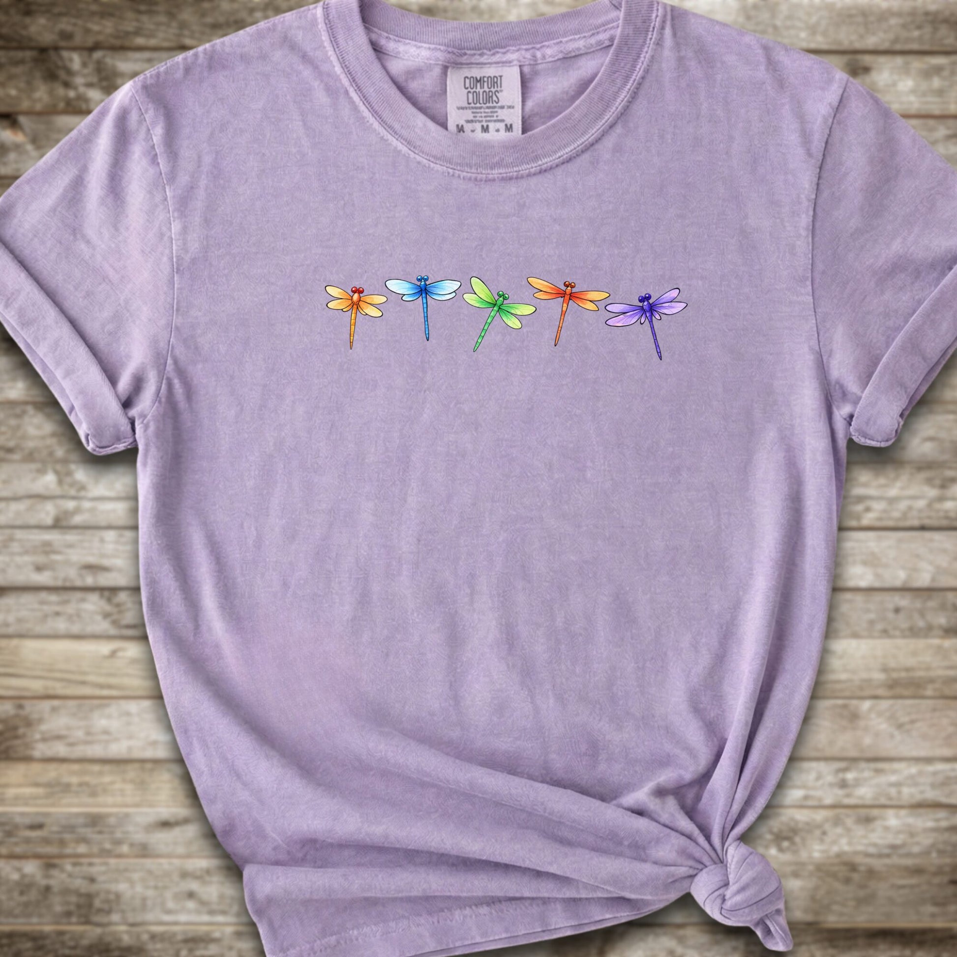 Purple short-sleeve t-shirt with a graphic of five colorful dragonflies across the chest, styled with a knot at the bottom hem and laid flat on a rustic wooden background.