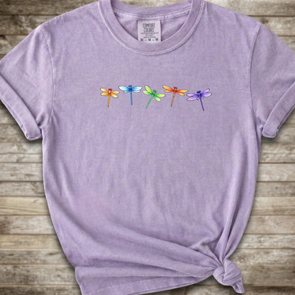 Purple short-sleeve t-shirt with a graphic of five colorful dragonflies across the chest, styled with a knot at the bottom hem and laid flat on a rustic wooden background.