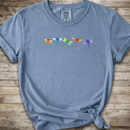 Blue short-sleeve t-shirt with a graphic of five colorful dragonflies across the chest, styled with a knot at the bottom hem and laid flat on a rustic wooden background.